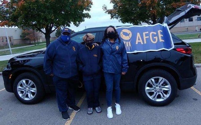 Three people standing in front of a SUV with an AFGE banner